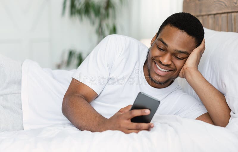 Homem Africano Utilizando Celular Navegando Na Internet Deitado Dentro Da Cama Foto de Stock - Imagem de jogo, descanso: 199568776