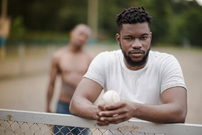 Homem afro-americano considerável com a bola do basebol que inclina-se na cerca na corte imagens de stock royalty free Homem afro-americano considerável com a bola do basebol que inclina-se na cerca na corte imagens de stock royalty free