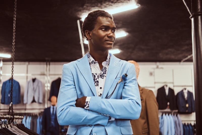 Homem afro-americano vestido no terno elegante azul que levanta com braços cruzados em uma loja clássica do menswear fotografia de stock Homem afro-americano vestido no terno elegante azul que levanta com braços cruzados em uma loja clássica do menswear fotografia de stock