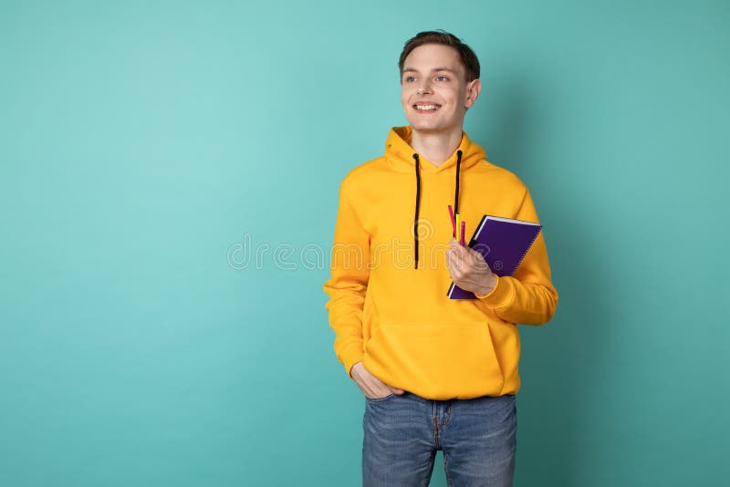 Homem alegre novo que veste o hoodie amarelo, guardando o caderno e a pena roxos Inspira??o a criar fotos de stock Homem alegre novo que veste o hoodie amarelo, guardando o caderno e a pena roxos Inspira??o a criar fotos de stock