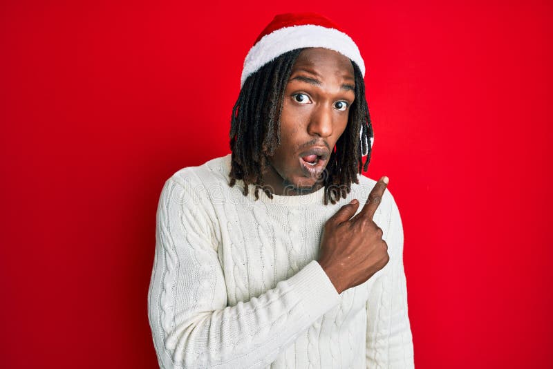 African american man with braids wearing christmas hat surprised pointing with finger to the side, open mouth amazed expression. African american man with braids wearing christmas hat surprised pointing with finger to the side, open mouth amazed expression