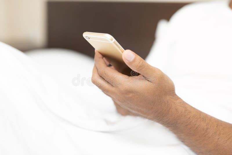 Homem ascendente próximo do viciado do telefone celular do homem novo da mão acordado na manhã na cama imagem de stock Homem ascendente próximo do viciado do telefone celular do homem novo da mão acordado na manhã na cama imagem de stock