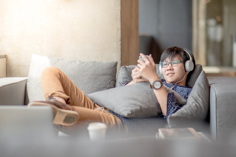 Homem asiático novo que usa o smartphone que relaxa no sofá fotos de stock Homem asiático novo que usa o smartphone que relaxa no sofá fotos de stock