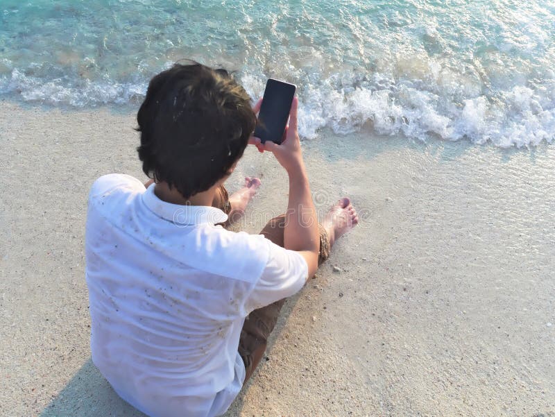 Homem asiático novo que usa o telefone esperto móvel no Sandy Beach Conceito das férias de verão imagens de stock royalty free Homem asiático novo que usa o telefone esperto móvel no Sandy Beach Conceito das férias de verão imagens de stock royalty free