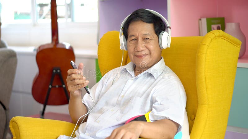 Homem asiático sênior usando smartphone escutando música com fones de ouvido, sentado no sofá em casa, relaxamento masculino