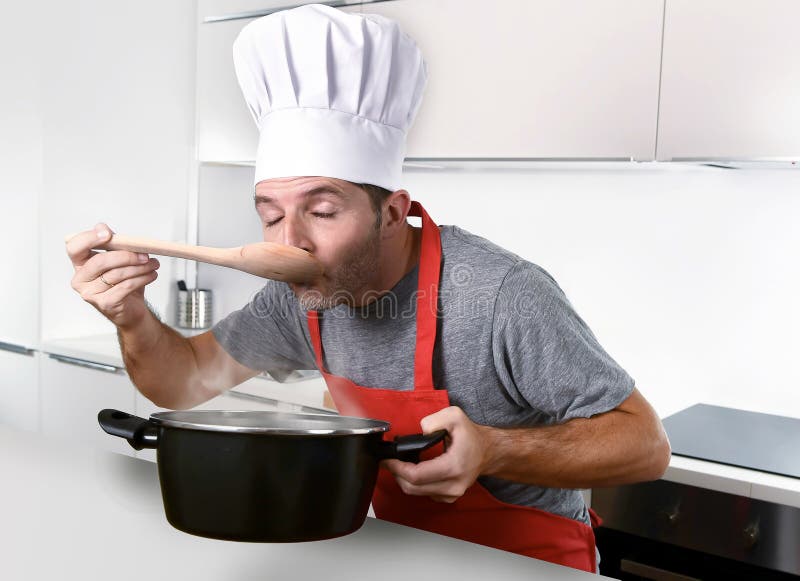 Homem atrativo novo no chapéu do cozinheiro chefe e no sabor vermelho dos testes do avental do cozinheiro imagem de stock royalty free Homem atrativo novo no chapéu do cozinheiro chefe e no sabor vermelho dos testes do avental do cozinheiro imagem de stock royalty free