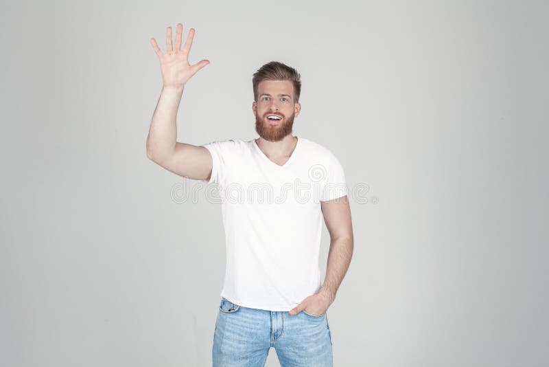 Homem bem sucedido com a barba lux?ria do gengibre acenando sua m?o na c?mera vestida em um t-shirt branco e na cal?as de ganga e fotografia de stock Homem bem sucedido com a barba lux?ria do gengibre acenando sua m?o na c?mera vestida em um t-shirt branco e na cal?as de ganga e fotografia de stock