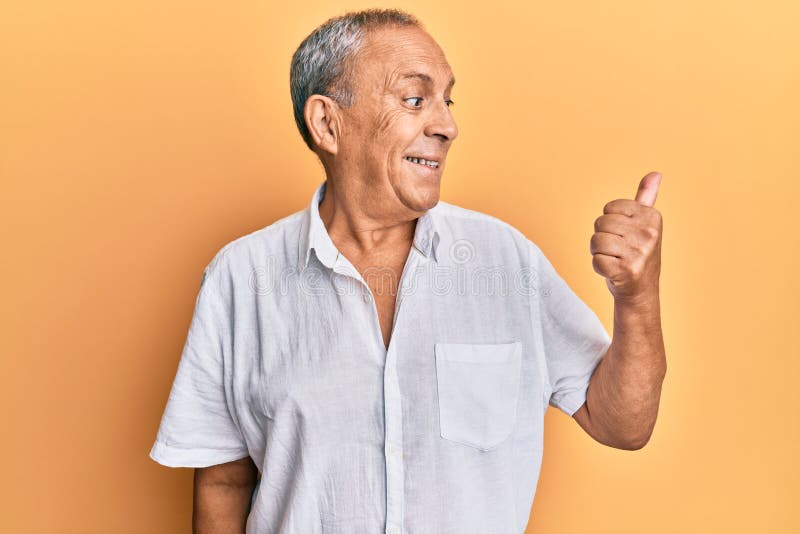 Handsome mature man wearing casual white shirt smiling with happy face looking and pointing to the side with thumb up. Handsome mature man wearing casual white shirt smiling with happy face looking and pointing to the side with thumb up