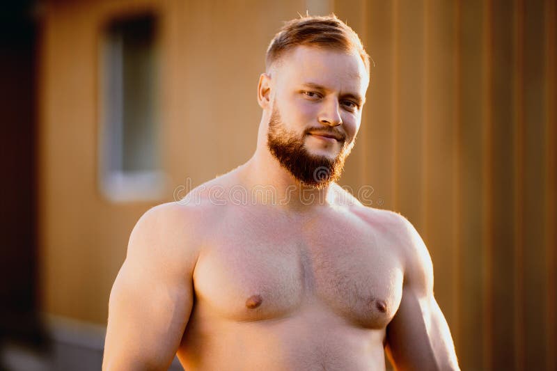 An athletic white man with beard stands alone near wooden rural house outdoors on summer evening in countryside. An athletic white man with beard stands alone near wooden rural house outdoors on summer evening in countryside.