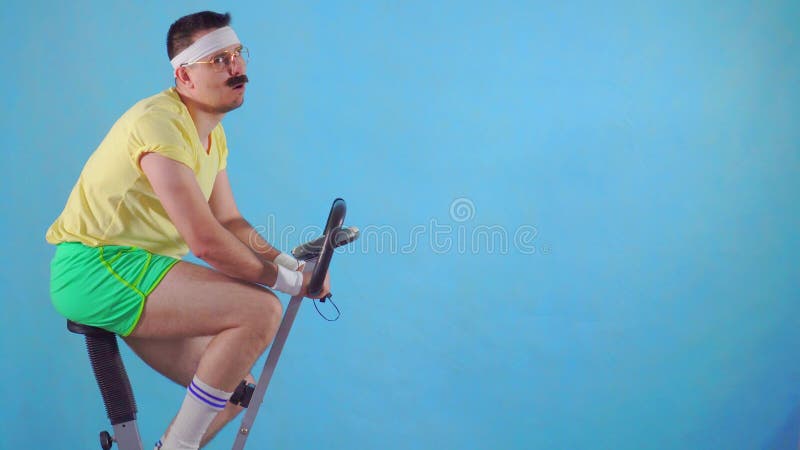 Homem cansado engraçado dos anos 80 com um bigode e vidros na bicicleta de exercício em um fundo azul