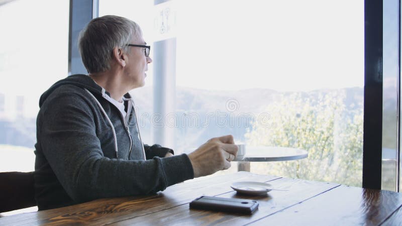 Homem caucasiano superior do estilo do esporte que olha para fora a janela no café Homem cinzento envelhecido que aprecia bebendo