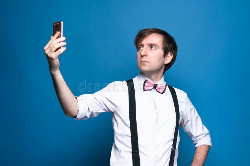 Homem com cabelo escuro em camisa cor-de-rosa com mangas enroladas, gravata-arco e espartilho preto tirando selfie sobre fundo az foto de stock Homem com cabelo escuro em camisa cor-de-rosa com mangas enroladas, gravata-arco e espartilho preto tirando selfie sobre fundo az foto de stock