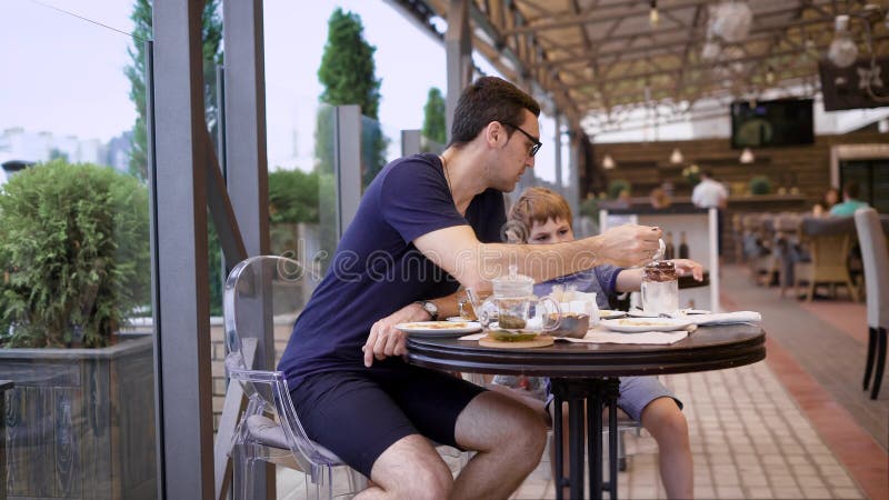 Homem considerável na camisa ocasional e nas culatras azuis que sentam-se no restaurante com filho e povos no fundo Pai novo