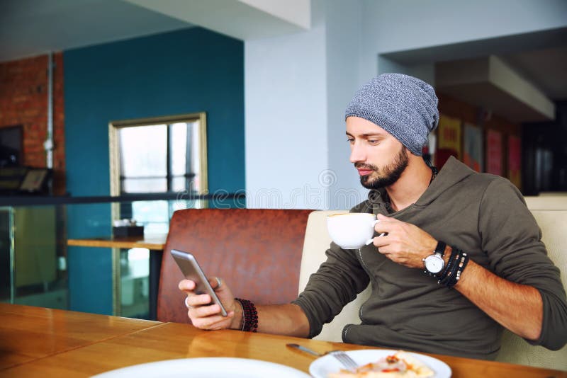 Homem considerável novo do moderno com a barba que senta-se no café usando um telefone celular, guardando a xícara de café imagens de stock royalty free Homem considerável novo do moderno com a barba que senta-se no café usando um telefone celular, guardando a xícara de café imagens de stock royalty free