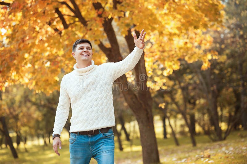 Homem considerável novo na camiseta e nas calças de brim que fica no parque alaranjado do outono que sorri e que acena a alguém fotografia de stock Homem considerável novo na camiseta e nas calças de brim que fica no parque alaranjado do outono que sorri e que acena a alguém fotografia de stock