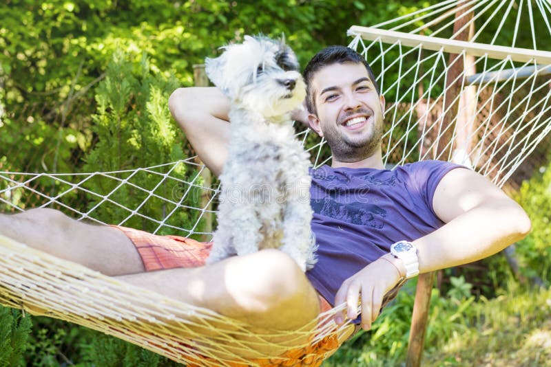Homem considerável novo que relaxa na rede com seu cão branco imagem de stock Homem considerável novo que relaxa na rede com seu cão branco imagem de stock
