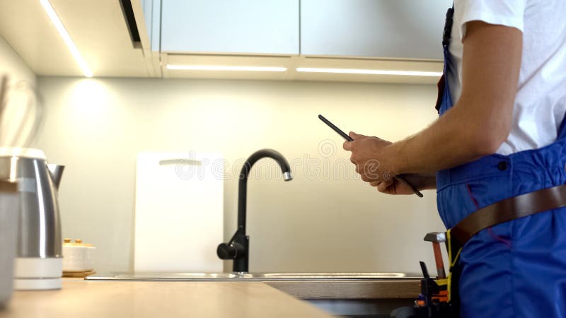 Homem de mão na torneira uniforme da cozinha, digitando relatório em tablet, encanamento foto de stock Homem de mão na torneira uniforme da cozinha, digitando relatório em tablet, encanamento foto de stock