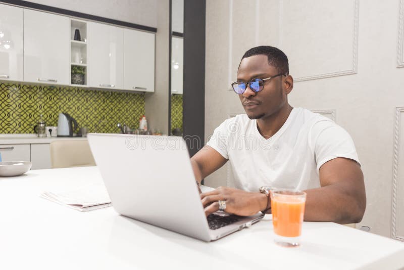 Homem de neg?cios afro-americano novo que trabalha em um port?til na cozinha em um interior moderno fotos de stock royalty free Homem de neg?cios afro-americano novo que trabalha em um port?til na cozinha em um interior moderno fotos de stock royalty free