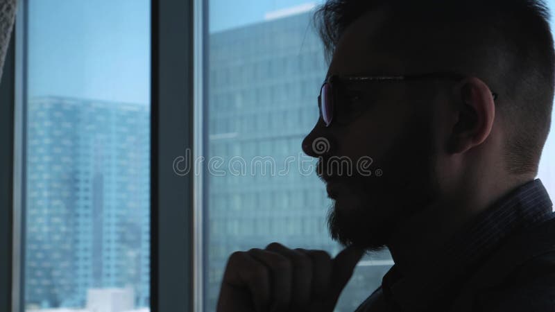 Homem de negócios bem sucedido novo com uma barba que pensa no fundo de uma janela que negligencia a baixa Negócio alto