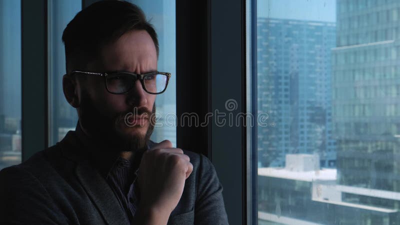 Homem de negócios bem sucedido novo com uma barba que pensa no fundo de uma janela que negligencia a baixa Negócio alto