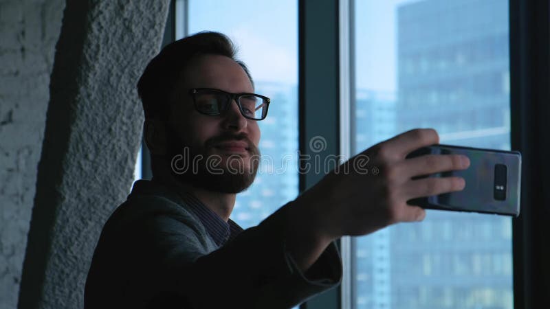 Homem de negócios bem sucedido novo que faz o selfie no fundo de uma janela que negligencia a baixa Edifício elevado do negócio