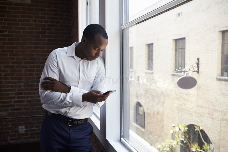 Homem de negócios Checking Phone Standing pela janela do escritório imagens de stock royalty free Homem de negócios Checking Phone Standing pela janela do escritório imagens de stock royalty free