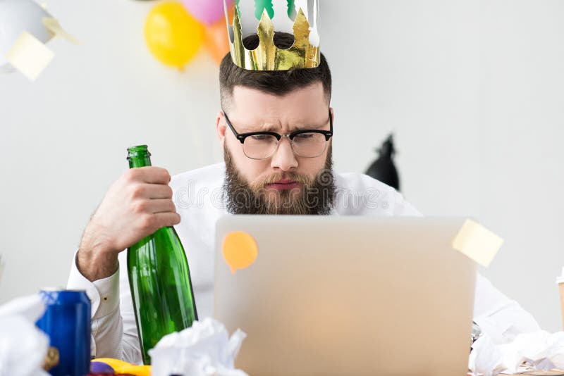 homem de negócios com a coroa de papel na cabeça e na garrafa do champanhe à disposição que olham a tela do portátil fotografia de stock homem de negócios com a coroa de papel na cabeça e na garrafa do champanhe à disposição que olham a tela do portátil fotografia de stock