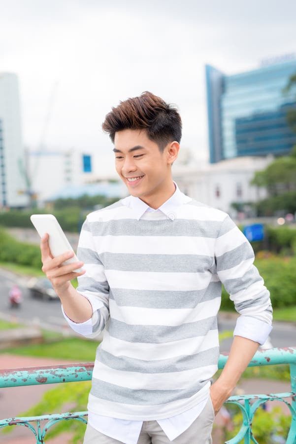 Homem de negócios considerável asiático novo que sorri ao ler seu smartphone Retrato da mensagem asiática da leitura do homem de imagem de stock Homem de negócios considerável asiático novo que sorri ao ler seu smartphone Retrato da mensagem asiática da leitura do homem de imagem de stock