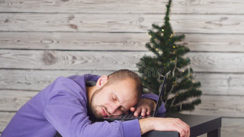 Homem de negócios novo cansado no escritório na véspera de ano novo Árvore de Natal no escritório Portátil na mesa Conceito do ne fotos de stock royalty free Homem de negócios novo cansado no escritório na véspera de ano novo Árvore de Natal no escritório Portátil na mesa Conceito do ne fotos de stock royalty free