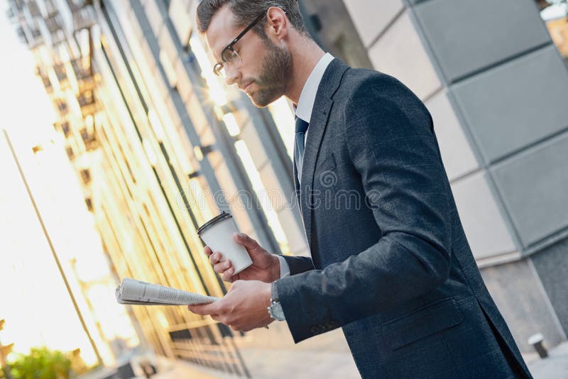 Homem de negócios novo no terno e vidros que guardam um copo de papel e que leem o jornal de negócio em suas mãos fotografia de stock Homem de negócios novo no terno e vidros que guardam um copo de papel e que leem o jornal de negócio em suas mãos fotografia de stock