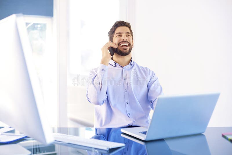 Homem de negócios novo que faz uma chamada ao trabalhar no portátil imagem de stock Homem de negócios novo que faz uma chamada ao trabalhar no portátil imagem de stock