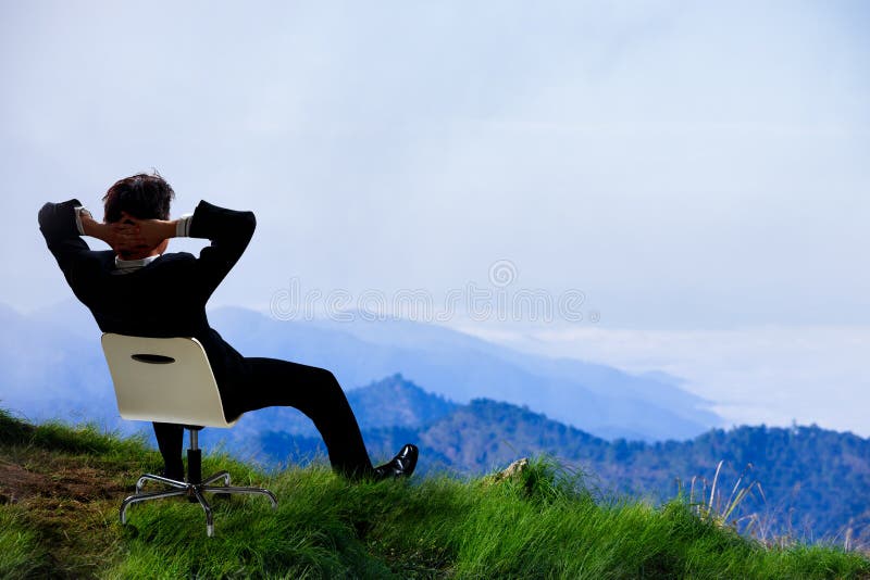 Homem de negócios novo que se senta em uma cadeira na parte superior da montanha fotografia de stock royalty free Homem de negócios novo que se senta em uma cadeira na parte superior da montanha fotografia de stock royalty free