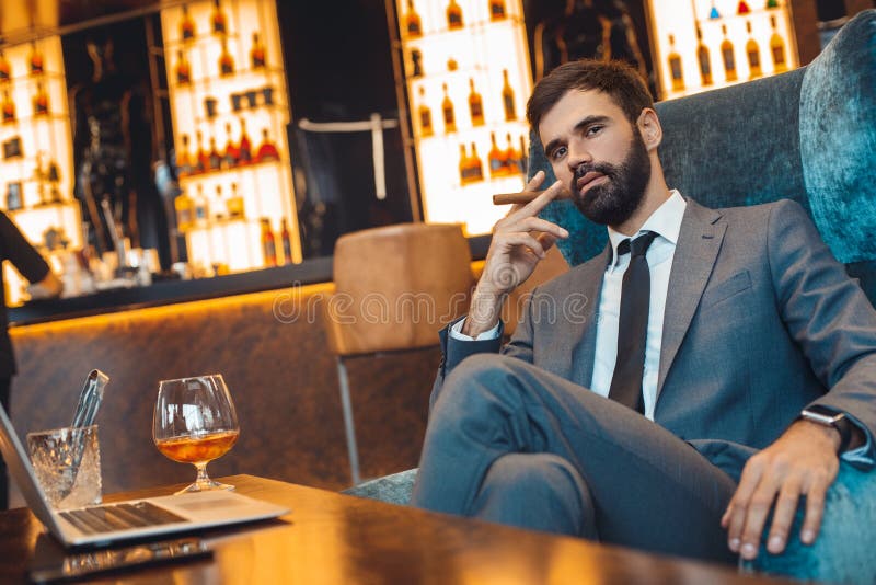 Homem de negócios que senta-se em um charuto de fumo da barra do centro de negócios imagem de stock royalty free Homem de negócios que senta-se em um charuto de fumo da barra do centro de negócios imagem de stock royalty free
