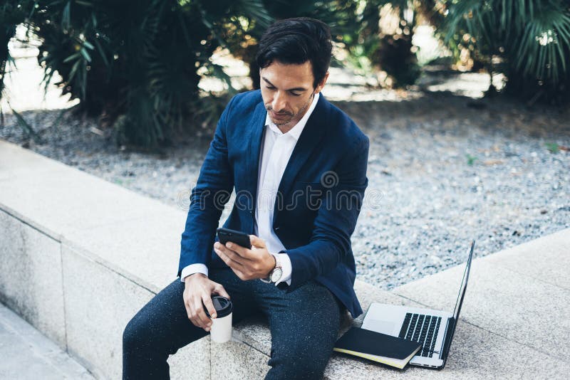 Homem de negócios de sorriso atrativo que usa o smartphone e o portátil contemporâneos ao tomar o resto durante o dia do trabalho foto de stock Homem de negócios de sorriso atrativo que usa o smartphone e o portátil contemporâneos ao tomar o resto durante o dia do trabalho foto de stock