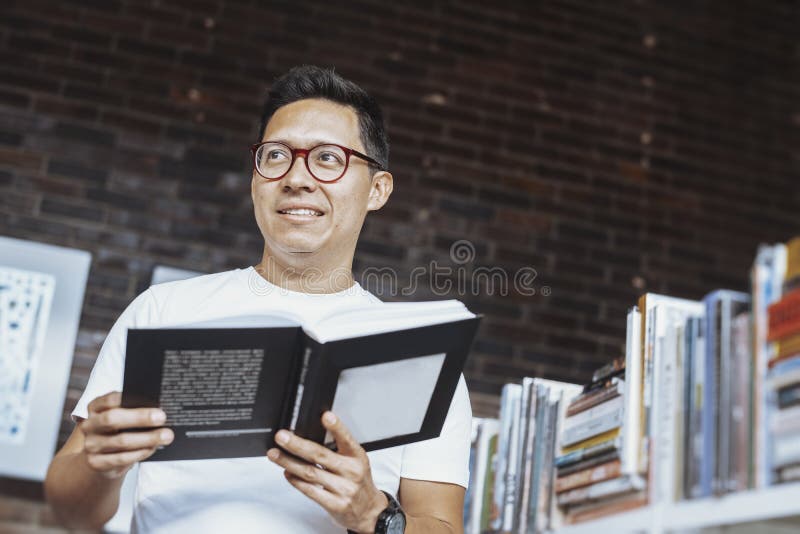 Homem de óculos pensativo novo que guarda o livro e que olha de lado fotos de stock Homem de óculos pensativo novo que guarda o livro e que olha de lado fotos de stock