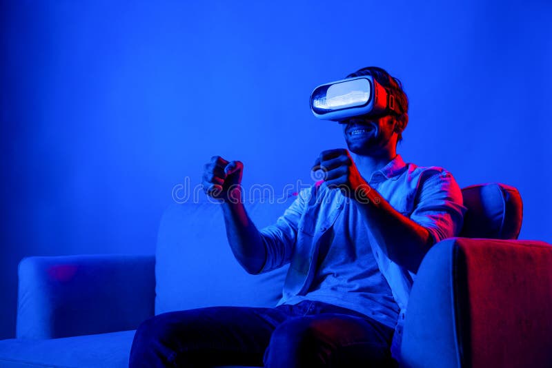 Happy man drive a car movement while wear VR glasses and sitting at sofa with neon light background. Smart gamer playing sport game while moving driving car gesture by using smart goggle. Deviation. Happy man drive a car movement while wear VR glasses and sitting at sofa with neon light background. Smart gamer playing sport game while moving driving car gesture by using smart goggle. Deviation.