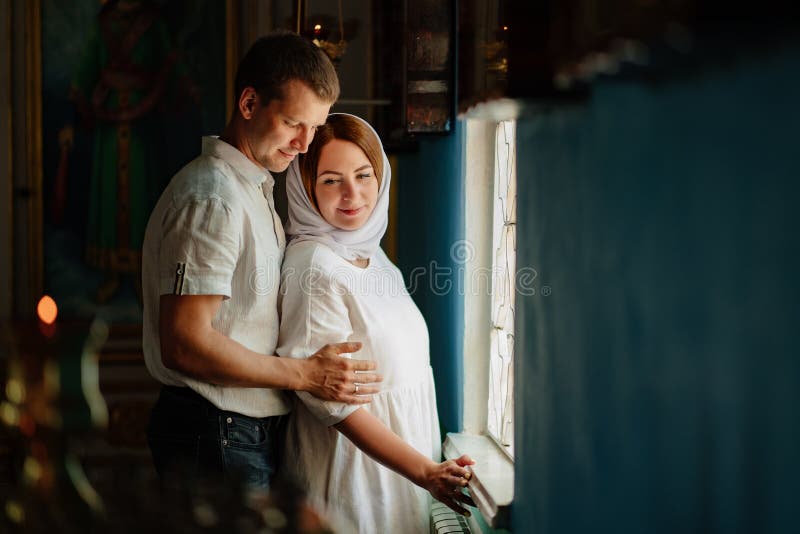 A men and a women in a headscarf and light-colored robes by the window in the church. baptism and wedding in the Orthodox Christian Church. family tradition to go to church for worship. A men and a women in a headscarf and light-colored robes by the window in the church. baptism and wedding in the Orthodox Christian Church. family tradition to go to church for worship.