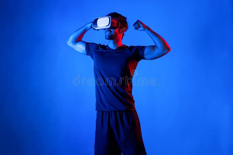 Happy man with VR goggle flexing muscle at neon light background. Healthy person with casual cloth relaxing and exercising by using virtual reality simulator technology. Represent healthy. Deviation. Happy man with VR goggle flexing muscle at neon light background. Healthy person with casual cloth relaxing and exercising by using virtual reality simulator technology. Represent healthy. Deviation.