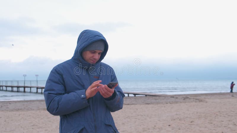 Homem em um azul abaixo do revestimento na praia da areia pelo mar que datilografa uma mensagem no telefone celular