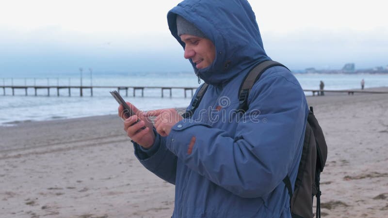 Homem em um azul abaixo do revestimento na praia da areia pelo mar que datilografa uma mensagem no telefone celular Vista lateral