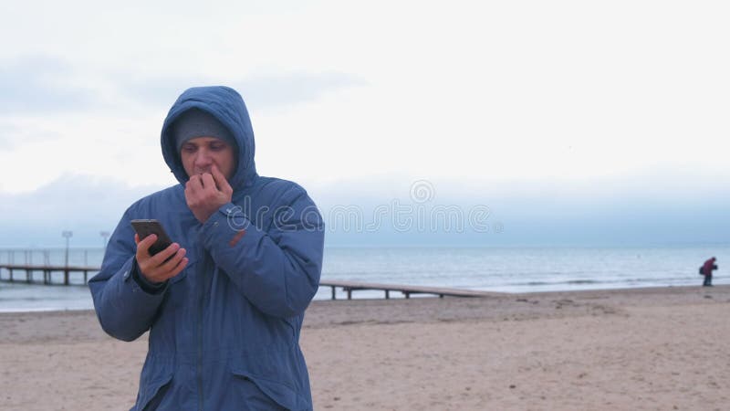 Homem em um azul abaixo do revestimento na praia da areia pelo mar que datilografa uma mensagem pelas mãos congeladas no telefone