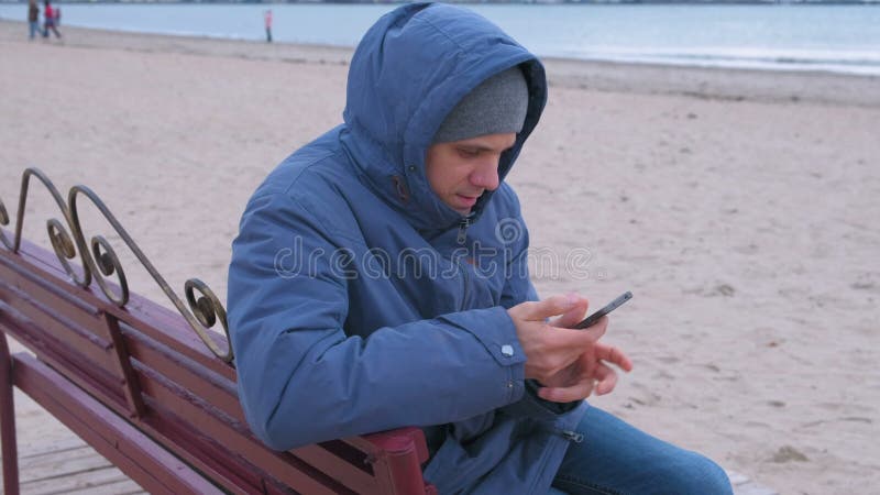 Homem em um azul abaixo do revestimento que senta-se em um banco na praia da areia e que datilografa uma mensagem no telefone cel
