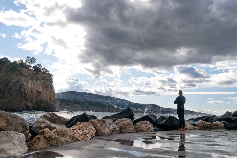 Homem em um terno molhado, surfista, estando na costa e olhando as ondas no fundo da montanha, Sorrento It?lia fotografia de stock Homem em um terno molhado, surfista, estando na costa e olhando as ondas no fundo da montanha, Sorrento It?lia fotografia de stock