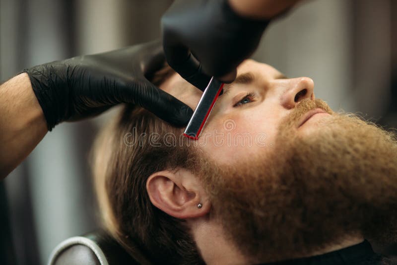 Homem farpado com a barba longa que obtém o cabelo à moda que barbeia, corte de cabelo, com a lâmina pelo barbeiro no barbeiro imagem de stock Homem farpado com a barba longa que obtém o cabelo à moda que barbeia, corte de cabelo, com a lâmina pelo barbeiro no barbeiro imagem de stock