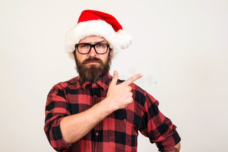 Handsome bearded man in Santa hat and glasses. Santa man pointing to the side over white wall. Christmas concept. Mock up, copy space for text. Hipster bearded man thinking for christmas gift ideas. Handsome bearded man in Santa hat and glasses. Santa man pointing to the side over white wall. Christmas concept. Mock up, copy space for text. Hipster bearded man thinking for christmas gift ideas