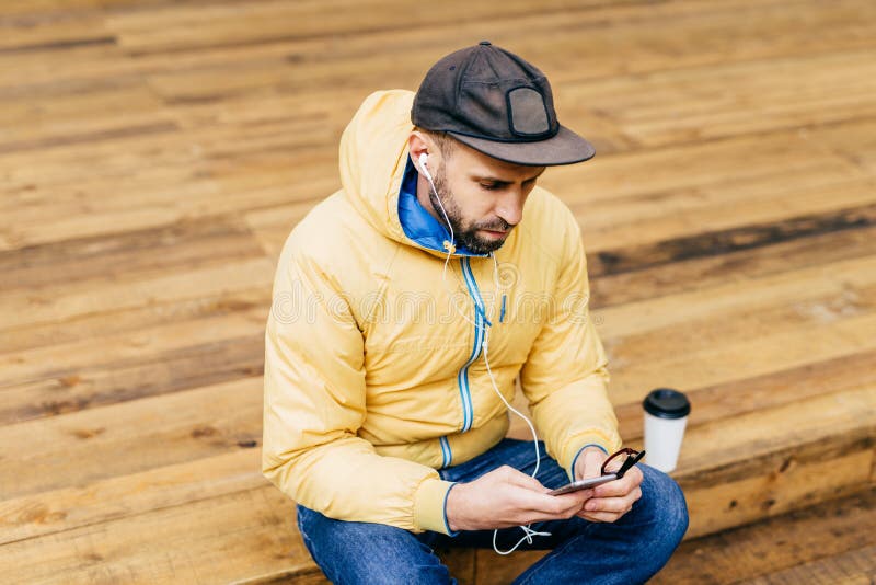 Homem farpado novo do moderno que veste o revestimento amarelo, o tampão preto à moda e as calças de brim sentando-se no assoalho fotos de stock Homem farpado novo do moderno que veste o revestimento amarelo, o tampão preto à moda e as calças de brim sentando-se no assoalho fotos de stock