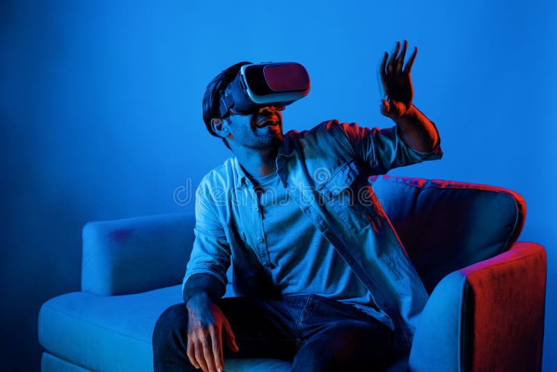 Happy man sitting at sofa while wearing VR goggle to watch funny movie. Caucasian person relaxed while looking at metaverse or virtual simulated world by using VR technology innovation. Deviation. Happy man sitting at sofa while wearing VR goggle to watch funny movie. Caucasian person relaxed while looking at metaverse or virtual simulated world by using VR technology innovation. Deviation.