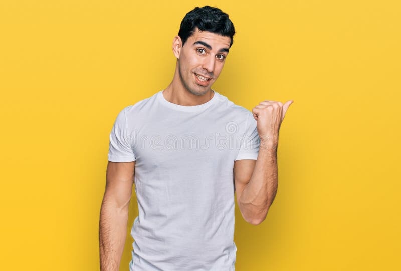 Handsome hispanic man wearing casual white t shirt smiling with happy face looking and pointing to the side with thumb up. Handsome hispanic man wearing casual white t shirt smiling with happy face looking and pointing to the side with thumb up