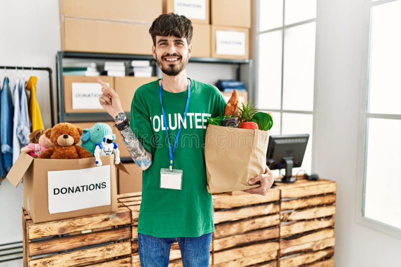 Hispanic man with beard wearing volunteer t shirt at donations point smiling happy pointing with hand and finger to the side. Hispanic man with beard wearing volunteer t shirt at donations point smiling happy pointing with hand and finger to the side