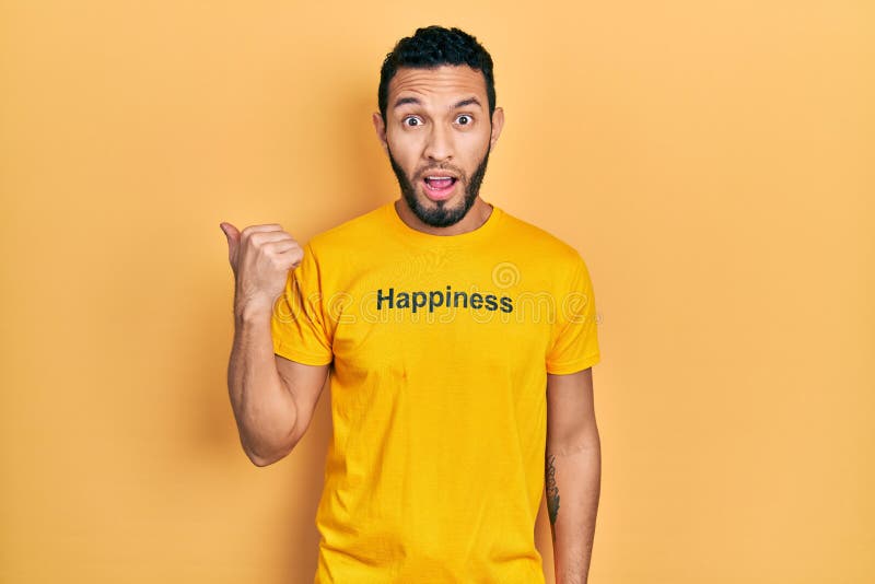 Hispanic man with beard wearing t shirt with happiness word message surprised pointing with hand finger to the side, open mouth amazed expression. Hispanic man with beard wearing t shirt with happiness word message surprised pointing with hand finger to the side, open mouth amazed expression